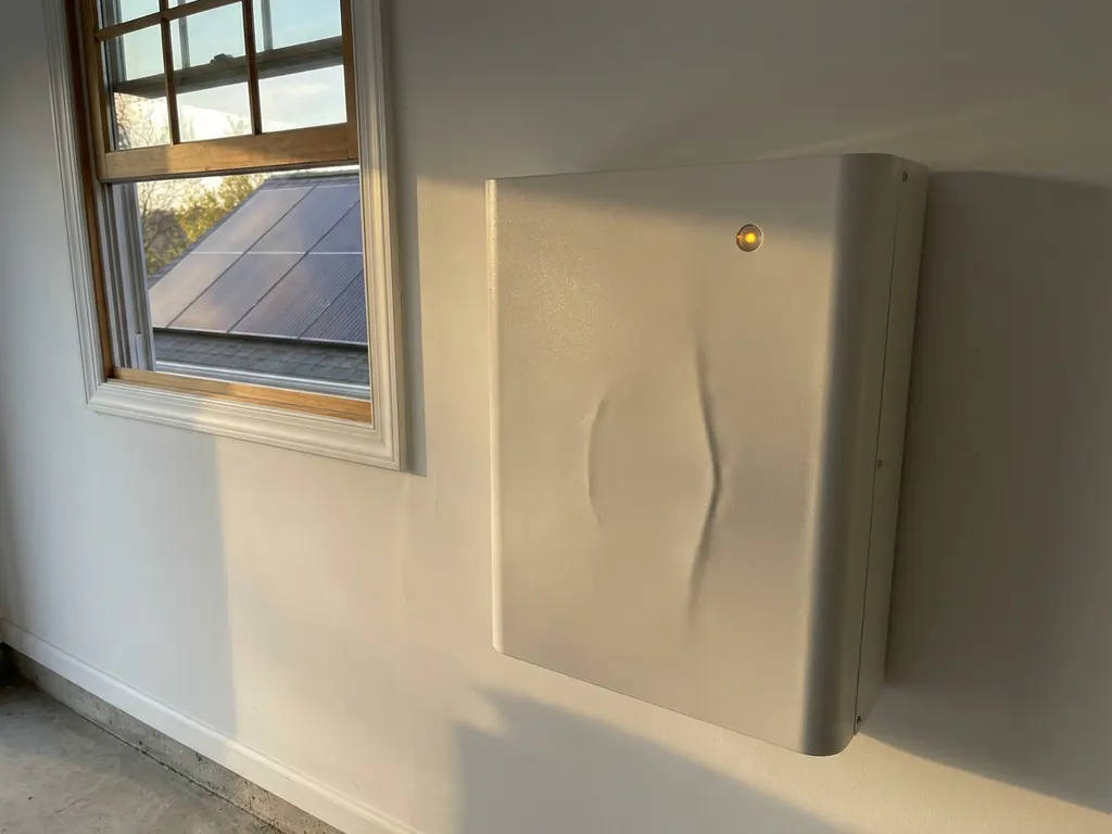 Swollen home battery unit on a white garage wall with amber indicator light, solar panel visible through sunlit window.